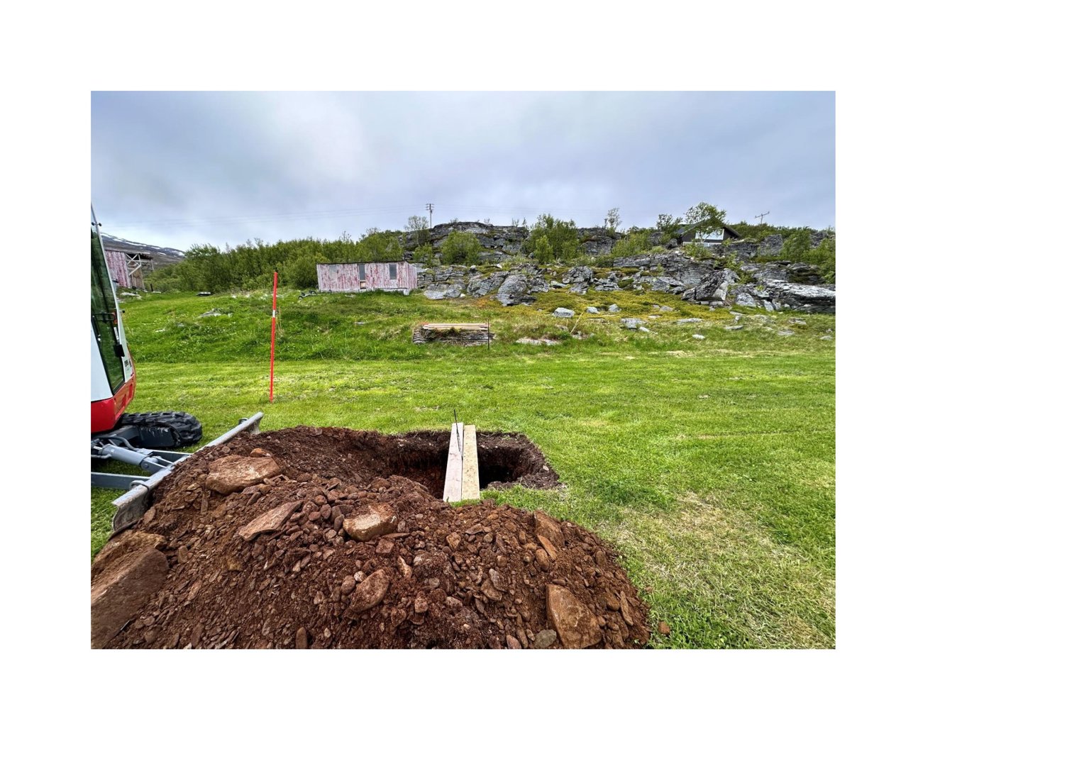 Fotografi av en byggeplass med utgravet grunn, en maskin og et bygg i bakgrunnen.