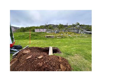 Fotografi av en byggeplass med utgravet grunn, en maskin og et bygg i bakgrunnen.
