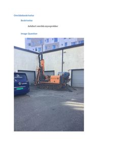 Fotografi av en byggeplass eller asfaltert område med en boremaskin og en bil parkert foran en bygning.