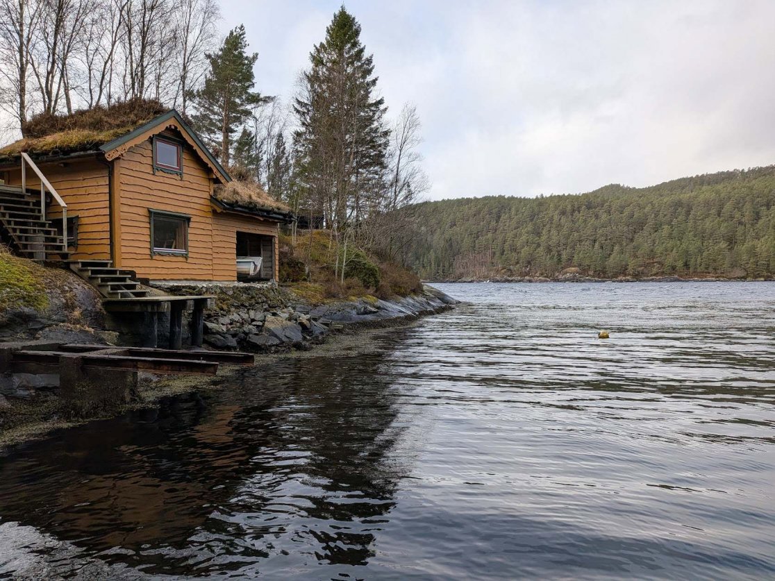 Fotografi av en liten hytte med grønt tak ved vannet, sett fra vannet mot land.