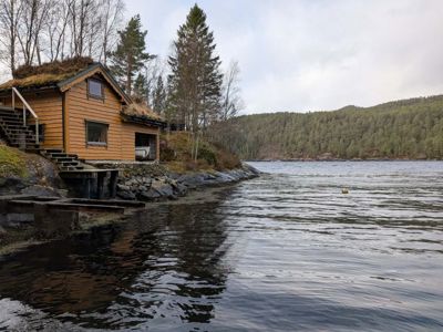 Fotografi av en liten hytte med grønt tak ved vannet, sett fra vannet mot land.