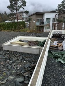 Fotografi av en byggeplass med en nybetont betongfundament eller kantmur i forgrunnen, og eksisterende boliger i bakgrunnen.
