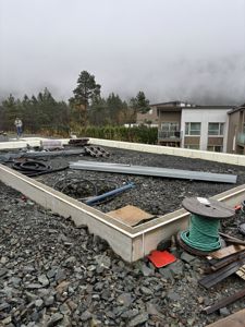 Fotografi av en byggeplass med en ny fundamentmur i forgrunnen, grus, byggematerialer og en person i bakgrunnen.