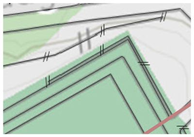 Dette bildet viser en detalj fra en situasjonsplan eller et tomtetegning. Det er synlig en grønn farget tomt med en svart kantlinje som definerer grensene. Det er også tegnet inn en vei eller gangvei (den hvite linjen med dobbeltstrekker) som krysser eller ligger ved siden av tomten. Konturene og linjene indikerer terrengform eller topografi.