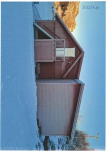 Fotografi av en rødbrun bygning med snø i forgrunnen og fjell i bakgrunnen.