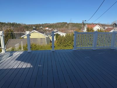 Fotografi av en terrasse eller balkong med mørke gulvplanker og hvit ramma. I bakgrunnen ser man nabolag med hus og trær under en klar blå himmel.