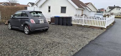 Fotografi av en bil parkert på en grusflate foran en hvit villa med hvit hekk og søppelcontainere.