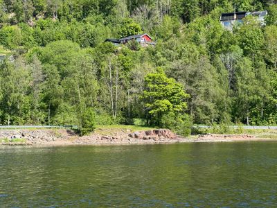 Fotografi av en kystlinje med vann i forgrunnen og en skogkledde bakke med hus i bakgrunnen.