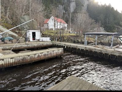 Fotografi av en kai med båter, et lite hvitt hus og en større bygning i bakgrunnen ved kysten.