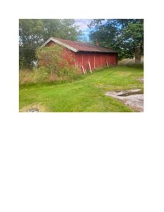 Fotografi av en rød, langbygget skur eller uthus sett fra siden, med grønt gress og trær i bakgrunnen.