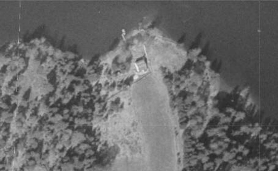 Et gammelt, svart-hvitt flyfoto som viser en bygning plassert på en halvøy eller landtunge, omgitt av vann og skog.