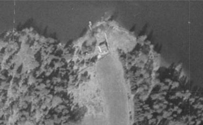 Et gammelt, svart-hvitt flyfoto som viser en bygning plassert på en halvøy eller landtunge, omgitt av vann og skog.