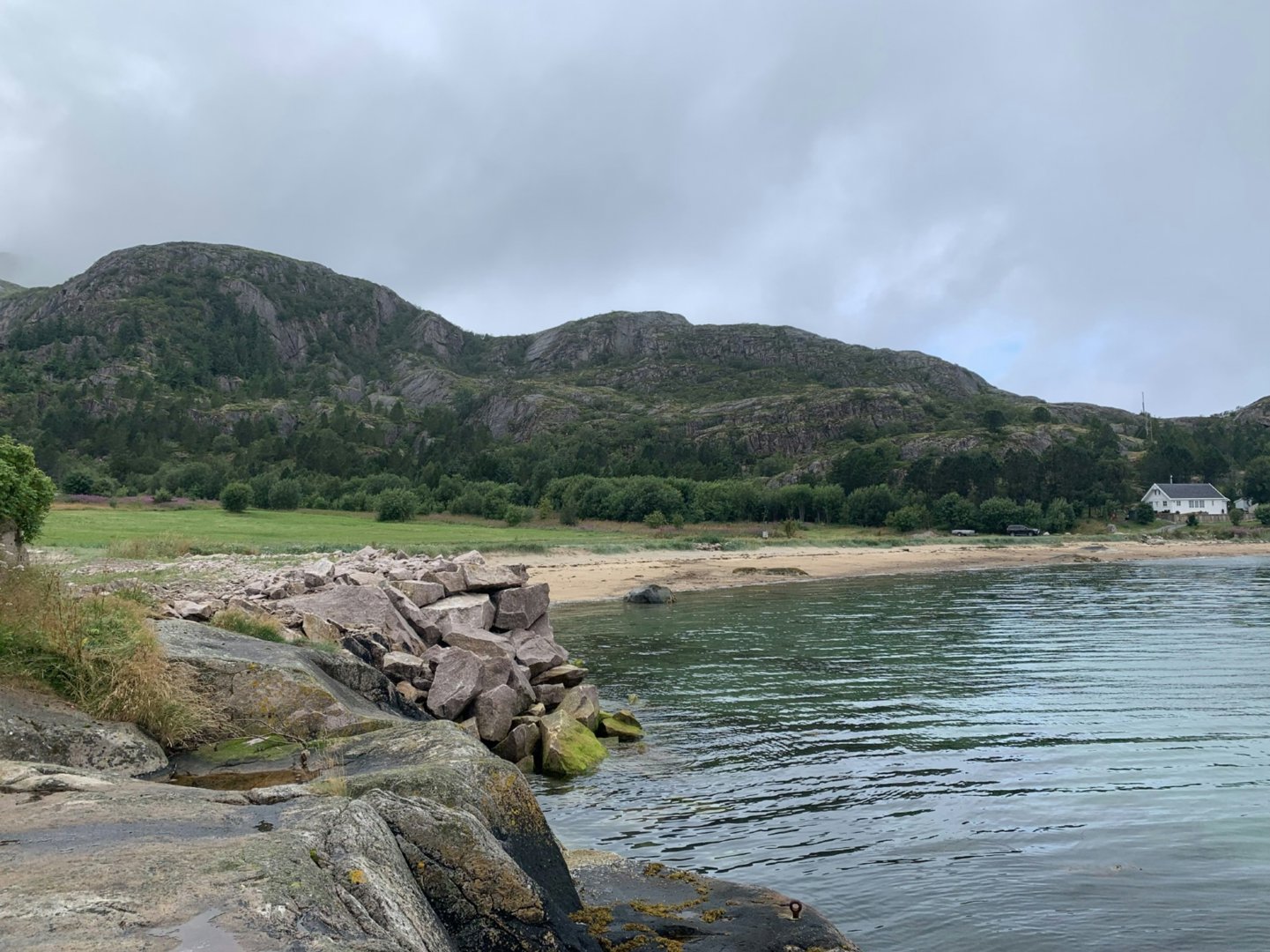 Fotografi av et kystlandskap med fjell, strand og vann. I bakgrunnen er det en hvit bygning (hus) synlig ved kanten av stranden.