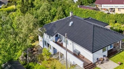 Fotografi av en hvit villa med mørk takflate sett fra luften, omgitt av grønt og naboer.