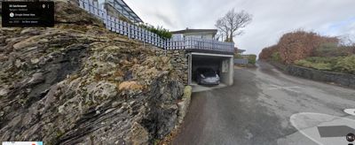 Fotografi av en bolig med garasje, sett fra en vei. Bildet viser en steinmur, en garasjeport med bil, og omgivelser.