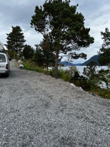 Fotografi av en grusvei som går langs en kystlinje med vann og fjell i bakgrunnen.