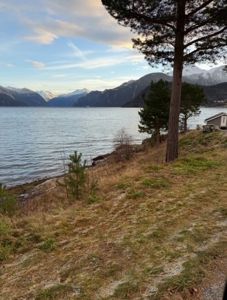 Fotografi av en utsikt over et vann med fjell i bakgrunnen, med en liten hytte synlig på høyre side.