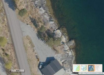 Flyfoto eller ortofoto av et område med en vei, en bygning, kystlinje og vann. Bildet inneholder også en målestokk og koordinatdata.