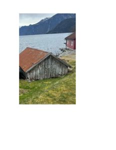 Fotografi av en gammel, forfallelig trebygning med rustet tak ved vannet, med en annen rød bygning og fjell i bakgrunnen.