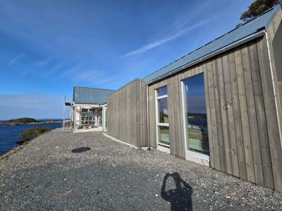 Fotografi av en moderne bygning med trefasade og metalltak, sett fra siden mot havet. Bildet viser også en skygge av fotografen og en del av en steinbelagt vei.