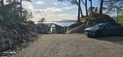 Fotografi av en moderne hytte eller villa plassert ved kysten, sett fra en grusvei med en bil i forgrunnen.