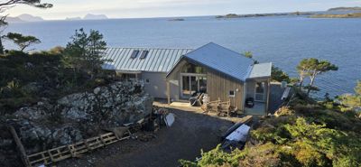 Fotografi av en moderne hytte eller villa som er under bygging eller renovering, sett fra et høydepunkt med utsikt over havet og øyer.