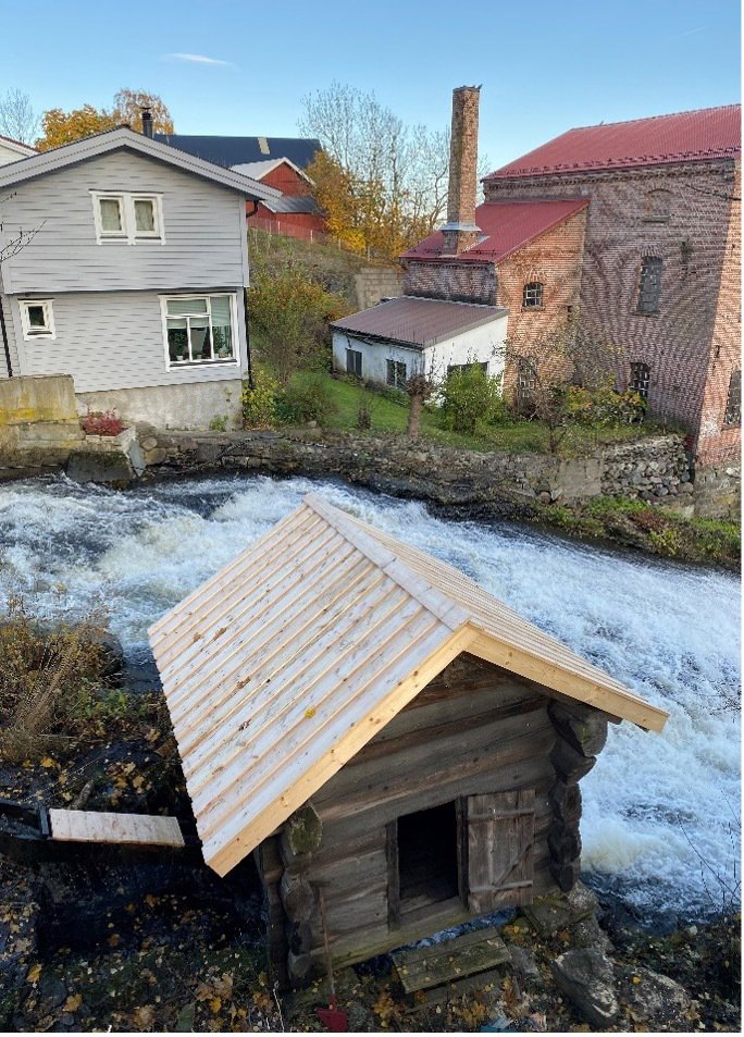 Fotografi av en liten hytte ved en elv, med andre bygninger i bakgrunnen.