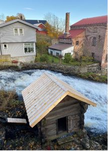 Fotografi av en liten hytte ved en elv, med andre bygninger i bakgrunnen.