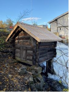Fotografi av en liten, gammel tømmerhytte eller utedo bygget på steiner ved en vannfall.