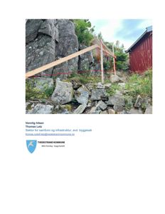 Fotografi av en steinmur med et trekonstruksjon (troll) og en rød bygning i bakgrunnen.