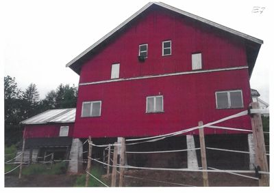 Fotografi av en rød, toetasjes bygning (muligens en uthus eller gård) sett fra siden. Bygningen er hevet på stolper, og det er en mindre tilbygning til venstre.