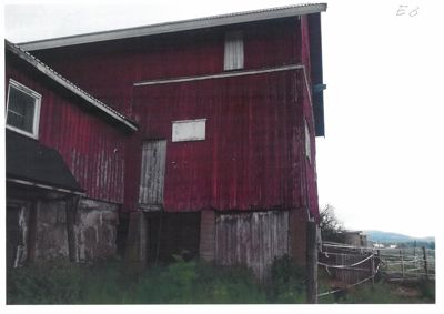 Fotografi av en rød, toetasje bygning (sannsynligvis en uthus eller gammel gård) sett fra siden. Bygningen har etasjeforskyvning og står på betongfundament.
