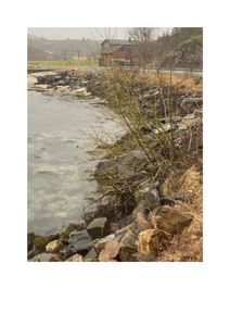 Fotografi av en kystlinje med steinmur, vann og en bygning i bakgrunnen.