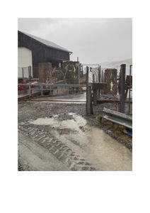 Fotografi av en bygning (gjerne en utesilo eller lagerhall) sett fra en grusvei eller kjørespor. Bildet viser en fuktig, regnig dag med synlige dype dyp i veien.