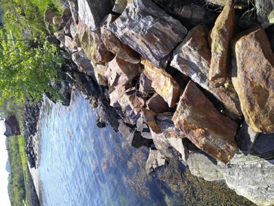 Fotografi av en steinmur langs et vassdrag med et rødt hus i bakgrunnen.