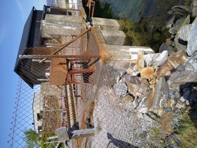 Fotografi av en bygning ved vannkanten med en rustet heis og en gjerde.