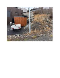 Fotografi av en bygning med mørk fasade og en mindre uthus, sett fra bakkenivå med en bakke i forgrunnen.