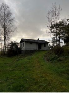 Fotografi av en hvit enebolig sett fra en skråning, omgitt av trær og grønt.