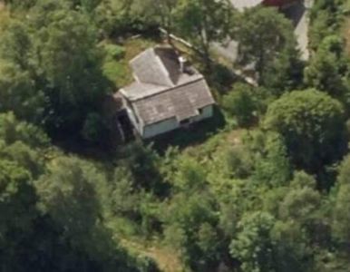 Luftfoto av en bygning (hus) sett fra oven, omgitt av skog og med en vei i bakgrunnen.