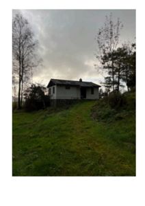 Fotografi av en liten hvit hytte eller hus sett fra en skråning, omgitt av trær og grønt.