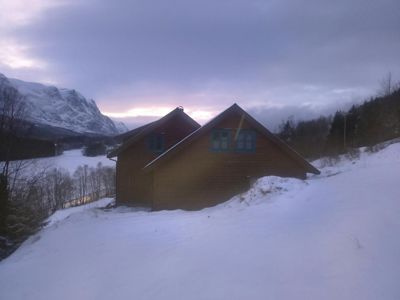 Fotografi av en hytte eller hus i snø, sett fra siden med fjell i bakgrunnen.