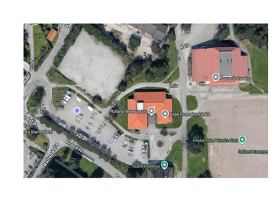 Satellittbilde (flyfoto) av et område med flere bygninger, parkeringsplasser og veier.