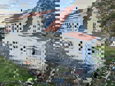 Fotografi av en hvit, toetasjes bygning med rødt tak som er under renovering eller nybygging. Bygningen er plassert på en grønn, kupert tomt med en steil fjellvegg i bakgrunnen. Det er synlig byggeutstyr, trelast og en rekkverk rundt en del av huset.