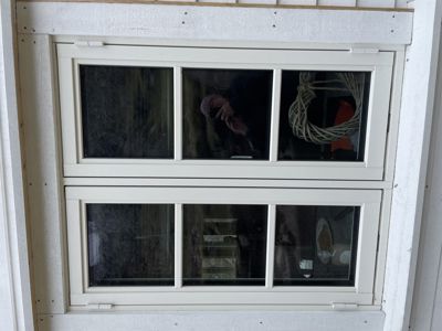 Fotografi av et hvitt, todelte vindu med tre glasspaneler i hver del, sett fra innsiden. I glasset er det synlig refleksjon av en person som tar bildet og gjenstander i rommet bak.