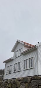 Fotografi av en hvit, trepanelert bygning sett fra nede. Bildet viser fasaden med vinduer, tak og en steinmur foran.