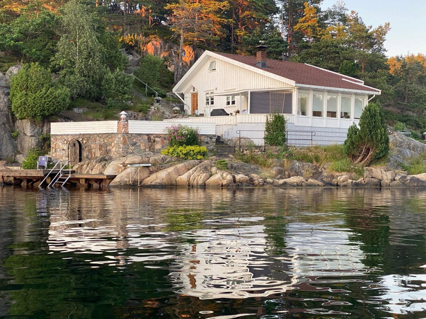 Fotografi av en hvit villa med steinmur ved kanten av vannet, sett fra vannet.