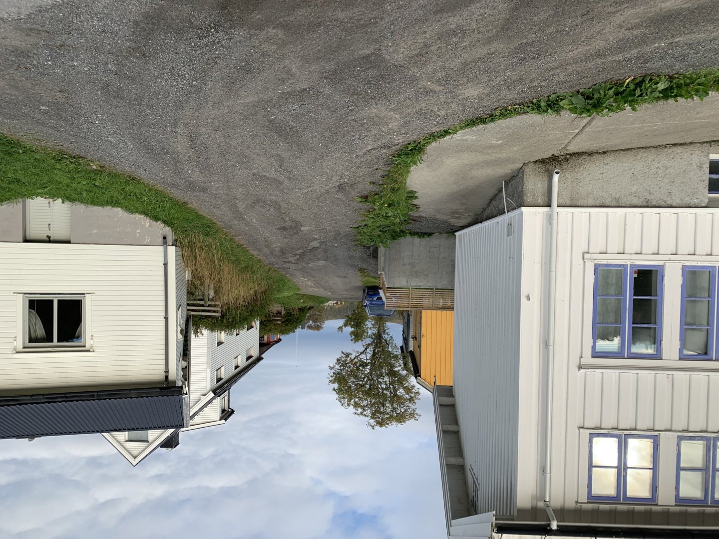 Fotografi av en bygning med hvit fasade og blå vindusrammer, sett fra siden. Bildet viser også en del av en vei, grus og en annen bygning i bakgrunnen.