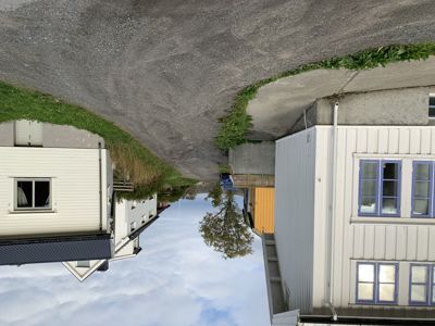 Fotografi av en bygning med hvit fasade og blå vindusrammer, sett fra siden. Bildet viser også en del av en vei, grus og en annen bygning i bakgrunnen.