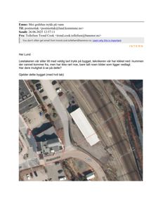 Satellittbilde (Google Maps) av et bygg med hvitt tak og omgivende infrastruktur, inkludert jernbanespor og vei.