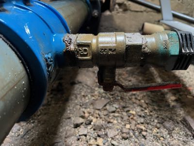 Fotografi av en blå vannmåler og en messingventil med en rød håndtak i en grusete underdel.